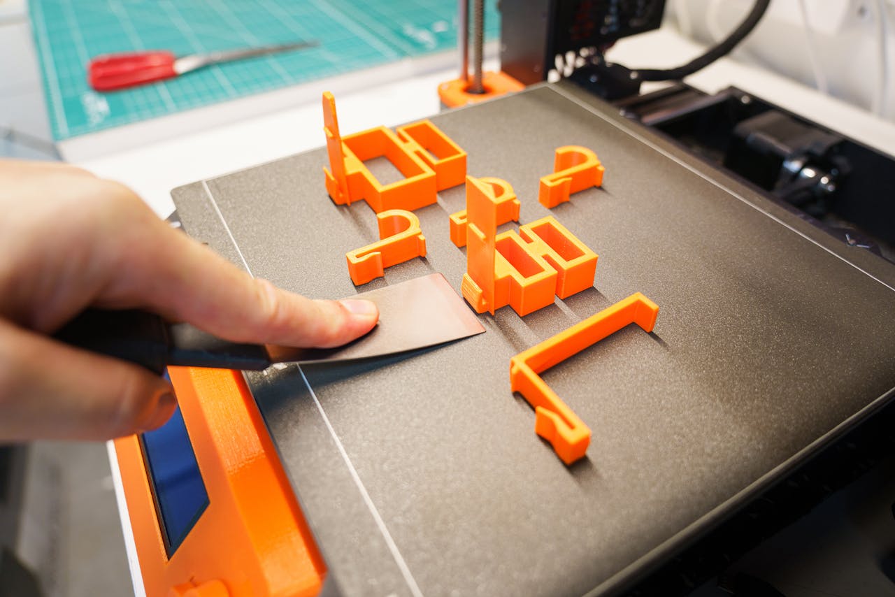 services-01 A hand carefully removes an orange 3D printed object from a printer bed using a spatula indoors.